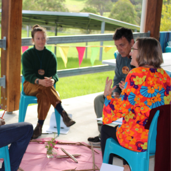 Small group seated in a circle on blue chairs, having a discussion in a covered outdoor space with colourful bunting.